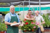 Jardinero senior utilizando portapapeles con clientes en el fondo del centro de jardinería - foto de stock
