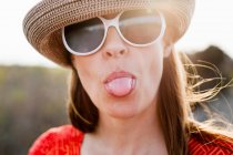 Mature woman wearing sunglasses and sunhat sticking out tongue — Stock Photo