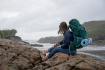 Mujer joven con mochila, sentada en roca, Constant Bay, Charleston, Isla Sur, Nueva Zelanda - foto de stock
