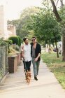 Молодая пара мужчин гуляет с собакой по пригородному тротуару — стоковое фото