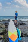 Молодий хлопчик стоїть на скелях біля моря, дивлячись на вигляд, дошка для серфінгу на передньому плані — стокове фото