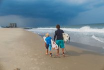 Vater und Sohn gehen am Strand entlang, tragen Surfbretter, Rückansicht — Stockfoto