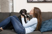Side view of young woman on sofa holding smartphone playing with dog — Stock Photo