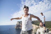 Young man wearing vest on cliff by ocean in arms open in jumping stance, Capo Caccia, Sardinia, Italy — Stock Photo