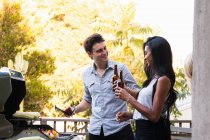 Young couple outdoors, holding beer bottles, cooking food on barbecue — Stock Photo