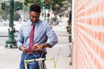 Young man standing beside bicycle, using smartphone, looking at wristwatch — Stock Photo
