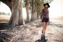 Молодая женщина в стиле бохо и шляпы прогуливаясь по обочине дороги — стоковое фото