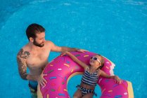 Chica joven en la piscina, relajándose en el anillo inflable, padre de pie junto a ella - foto de stock