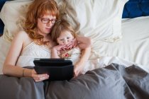 Мать и дочь лежат в постели, смотрят на цифровой планшет — стоковое фото