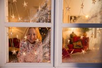 Девушка смотрит в окно с рождественскими украшениями — стоковое фото