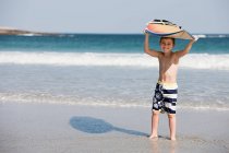 Мальчик держит доску для серфинга над головой на берегу моря — стоковое фото