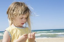 Ragazza con i capelli biondi flyaway guardando conchiglia sulla spiaggia — Foto stock