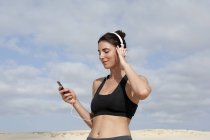 Mulher adulta média selecionando música para fones de ouvido enquanto se exercita na praia — Fotografia de Stock