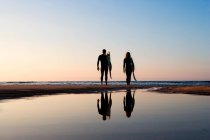 Vista posteriore della coppia in piedi sulla spiaggia, riflessione in acqua — Foto stock