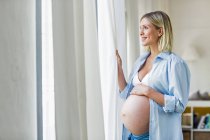 Повна вагітність молода жінка дивиться з вікна — стокове фото