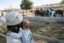 Mãe segurando filho jovem ao ar livre, assistindo cavalos na fazenda, visão traseira — Fotografia de Stock