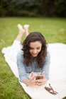 Молода жінка лежить в парку, дивлячись на мобільний телефон, посміхаючись — стокове фото