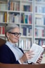 Седовласая взрослая женщина, читающая книгу с книжных полок — стоковое фото