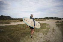 Donna anziana che cammina lungo la spiaggia, portando la tavola da surf, vista posteriore — Foto stock