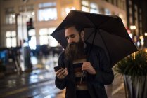 Hombre caminando en la ciudad por la noche, usando paraguas, mirando el teléfono inteligente, Centro, San Francisco, California, EE.UU. - foto de stock