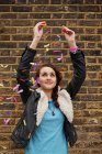 Young woman dropping colourful confetti against brick wall — Stock Photo