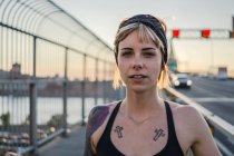 Young tattooed woman running on bridge taking a break with sunset behind — Stock Photo