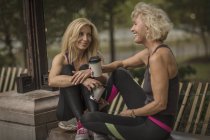 Deux amies mûres s'entraînent dans le parc, assises sur un mur avec du café à emporter — Photo de stock