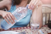 Обрезанный снимок зрелой женщины, наливающей стакан бутилированной воды за стол — стоковое фото