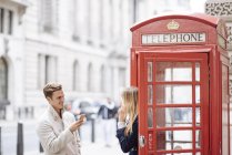 Pareja joven con teléfono inteligente junto a la cabina del teléfono rojo, Londres, Inglaterra, Reino Unido - foto de stock