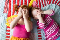 Портрет двух юных сестёр, лежащих на одеяле, покрывающих лицо руками — стоковое фото