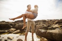 Молодий чоловік носить дівчину через кам'яний басейн на пляжі — стокове фото