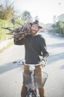 Young man carrying bunch of sticks on shoulder on bike — Stock Photo