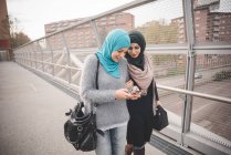Two female friends on footbridge reading smartphone texts — Stock Photo