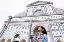 Low angle view of young woman using digital camera in front of church looking up, Piazza Santa Maria Novella, Florence, Tuscany, Italy — Stock Photo