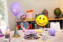 Two girls sitting at birthday party table with cake playing with smiley face balloon — Stock Photo