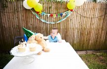 Niño sentado en la mesa con juguetes suaves con sombrero de fiesta - foto de stock