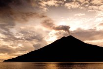Erupting volcano silhouette above water with cloudy sky — Stock Photo