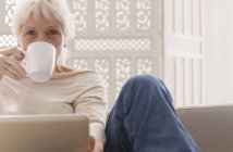 Mujer mayor bebiendo café y trabajando en el ordenador portátil - foto de stock