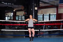 Portrait of female boxer leaning against boxing ring ropes, full length — Stock Photo
