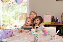 Tres niños sentados en la mesa de cumpleaños abrazándose - foto de stock
