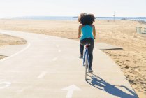 Mid adult woman cycling on pathway at beach, rear view — Stock Photo