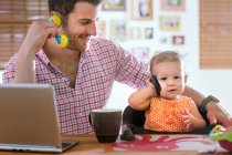 Hombre y bebé sentados en el mostrador de cocina jugando con teléfono inteligente y teléfono de juguete - foto de stock