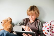 Jeune garçon utilisant une tablette numérique tout en étant assis sur son lit — Photo de stock