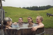 Cuatro amigos adultos riendo mientras se relajan en la bañera de hidromasaje rural, Sattelbergalm, Tirol, Austria - foto de stock