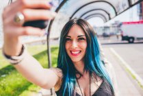 Jeune femme avec des cheveux bleus teints trempés prenant smartphone dans un abri de bus urbain — Photo de stock