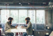 Взрослые хипстеры-близнецы сидят лицом к лицу за столом в офисе — стоковое фото