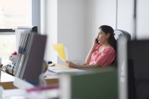Mujer sosteniendo papeles y sentada en la mesa de trabajo y hablando por teléfono móvil - foto de stock