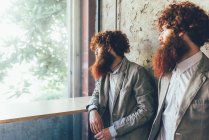 Identical male hipster twins looking out of office window — Stock Photo
