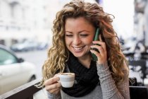 Mujer en la cafetería sosteniendo taza de café haciendo llamada telefónica sonriendo - foto de stock