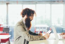 Jovem hipster masculino com cabelo encaracolado e barba usando tablet digital na mesa — Fotografia de Stock